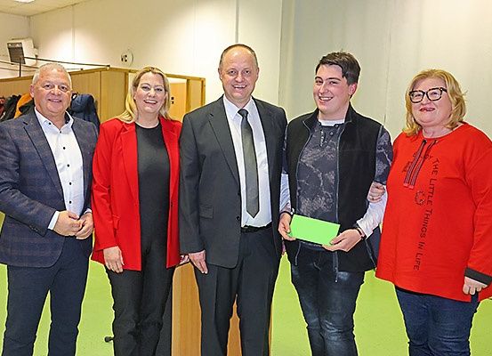 Fünf Menschen stehen lächelnd zusammen, ein Mann hält eine grüne Mappe, vor einer weißen Wand und einem Holzgestell.