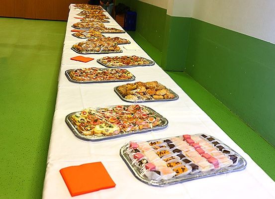 Ein langer Tisch ist mit mehreren Tabletts mit Essen gedeckt, darunter Sandwiches, Gebäck und andere Gerichte, die auf einer weißen Tischdecke platziert sind. Der Hintergrund zeigt grüne und weiße Wände.