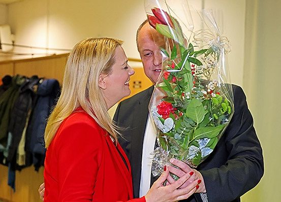Ein Mann in einem Anzug hält einen Blumenstrauß, während eine Frau in einem roten Blazer ihn anlächelt. Sie stehen drinnen in der Nähe einer Wand mit aufgehängten Jacken.