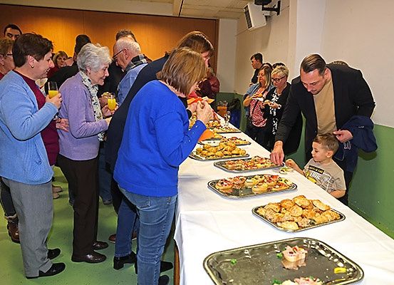 Mehrere Menschen stehen um einen Buffettisch und essen. Einige halten Gläser mit Orangensaft. Ein Mann füttert einen kleinen Jungen.