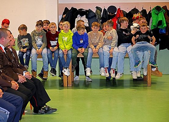 Eine Gruppe von Kindern sitzt auf einer Bank, mit einem Mann in Uniform vor ihnen. Der Raum hat grünen Boden und eine Wand mit hängenden Jacken.