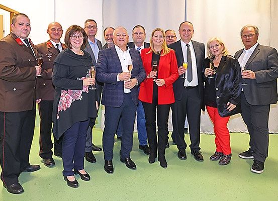 Eine Gruppe formell gekleideter Personen, einschließlich Männer und Frauen, stehen eng beieinander, lächeln und halten Weingläser. Sie scheinen ein Ereignis zu feiern.