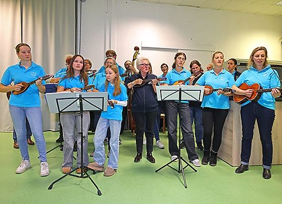 Eine Gruppe von Frauen in blauen Shirts und Jeans spielt Mandolinen, während andere zusehen. Sie befinden sich in einem Raum mit grünem Boden und weißen Wänden.