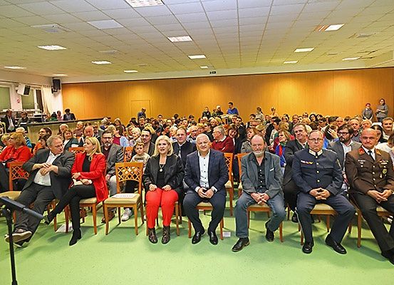 Eine Gruppe von Personen, einschließlich formeller und militärischer Mitarbeiter, sitzt in Stühlen in einem Raum. Einige tragen Brillen und halten Bücher.