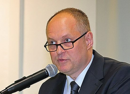 Ein Mann in Anzug und Krawatte spricht in ein Mikrofon in einem Raum. Er trägt eine Brille und sieht ernst aus. Das Mikrofon ist schwarz und silber.