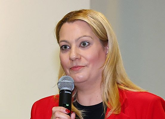 Eine Frau mit blonden Haaren, die einen roten Blazer trägt und ein Mikrofon hält, spricht möglicherweise bei einer Veranstaltung.