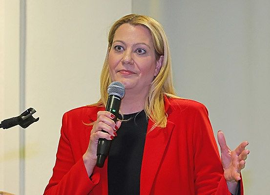 Eine Frau in einem roten Blazer spricht in ein Mikrofon in einem Konferenzraum. Sie lächelt und gestikuliert mit ihren Händen.