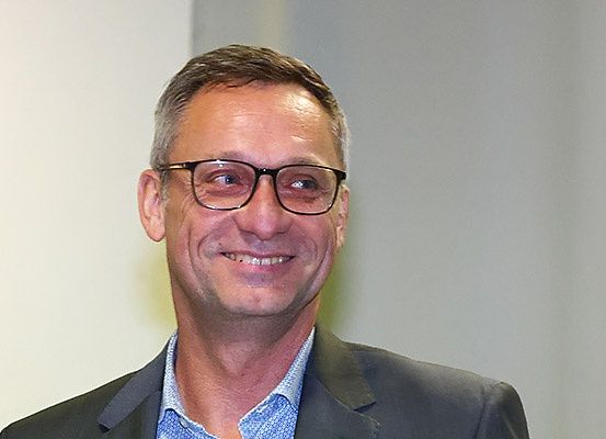 Ein Mann mit Brille und blauem Hemd, der steht und lächelt, ist in einem schwarzen Anzug gekleidet.
