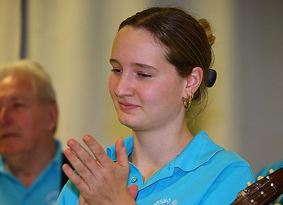 Eine Frau mit braunen Haaren in einem Pferdeschwanz klatscht mit den Händen und trägt ein blaues Poloshirt mit Logo auf der linken Seite. Sie lächelt und schaut nach vorne. Hinter ihr sieht man verschwommen einen Mann, der sie ansieht und ebenfalls ein blaues Poloshirt trägt.