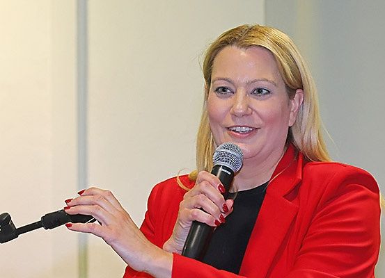 Eine Frau mit blonden Haaren lächelt und hält ein Mikrofon in der Hand, gekleidet in einem roten Blazer. Sie scheint auf einer Konferenz zu sprechen.