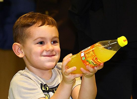 Ein kleiner Junge hält eine Flasche Orangensaft und lächelt, mit zwei Personen im Hintergrund.