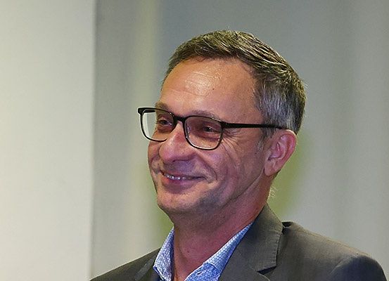Ein Mann mit kurzen, gepflegten Haaren, der eine Brille und einen Anzug trägt, lächelt in die Kamera. Er scheint sich in einem professionellen Umfeld zu befinden.