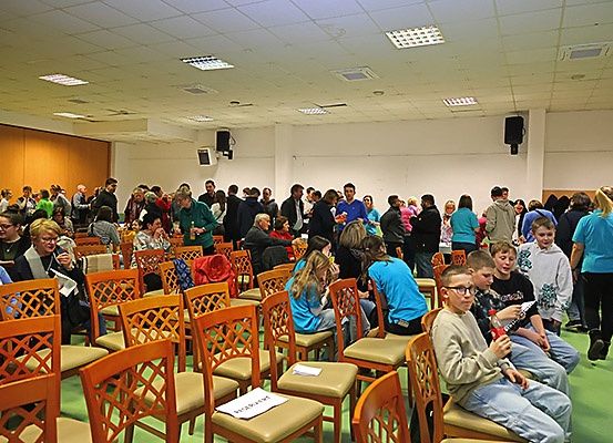 Eine große Indoor-Versammlung mit Menschen, die auf Stühlen sitzen, einige stehen und andere gehen herum. Einige Personen halten Getränke in der Hand. Der Raum hat weiße Wände, eine Decke mit Lichtern und einige an den Wänden montierte Lautsprecher.