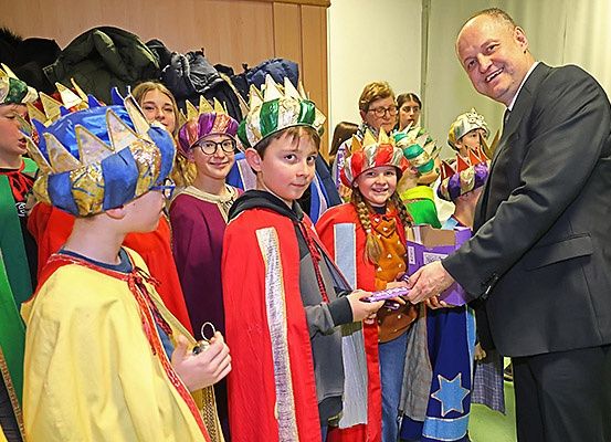 Ein Mann in einem Anzug verteilt Süßigkeiten an Kinder in bunten Kostümen, wahrscheinlich während eines festlichen Ereignisses.
