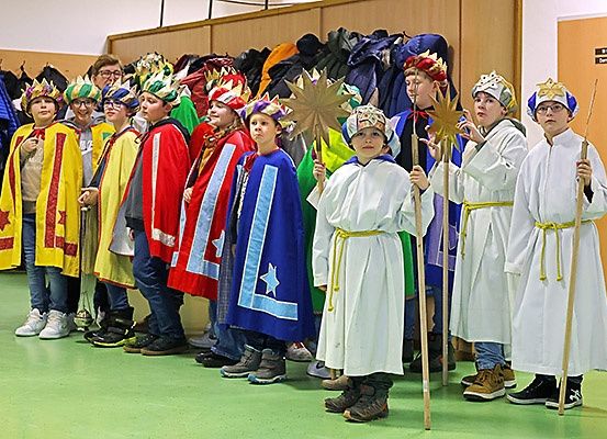 Eine Gruppe von Kindern in farbenfrohen Kostümen und mit Requisiten, möglicherweise für ein Theaterstück, steht in einer Reihe. Einige tragen Kronen.
