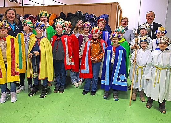 Eine Gruppe von Kindern in farbenfrohen Kostümen und Kronen steht zusammen in einem Raum und lächelt für ein Foto. Sie tragen verschiedenfarbige Umhänge und einige halten Requisiten. Hinter ihnen sind auch Erwachsene anwesend.