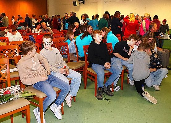 Eine Gruppe von Menschen sitzt in Stühlen in einem Raum. Einige tragen Brillen. Eine Blume ist auf einem Stuhl platziert. Der Raum ist schwach beleuchtet.