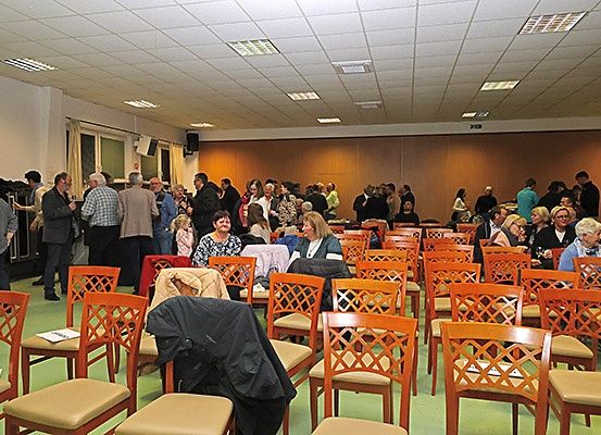 Ein großer Raum mit vielen versammelten Menschen, einige stehend, andere auf Stühlen sitzend, mit einem Tisch, der mit weißem Stoff bedeckt ist, einem Lautsprecher und einer offenen Tür auf der linken Seite.