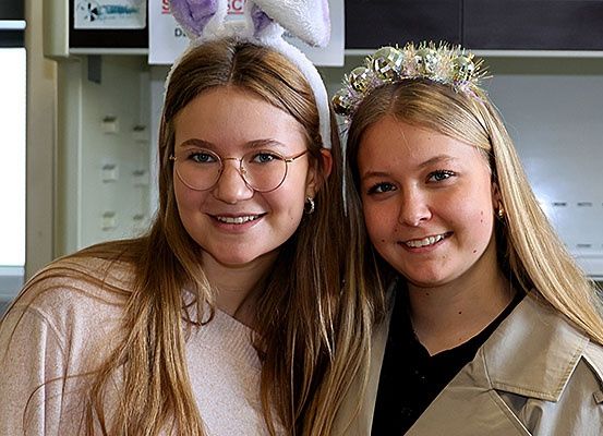 Zwei junge Frauen mit festlichen Haarbändern, eine mit Hasenohren und die andere mit einer glitzernden Krone, lächeln für die Kamera. Sie stehen nah beieinander in einem Raum mit weißen Wänden.