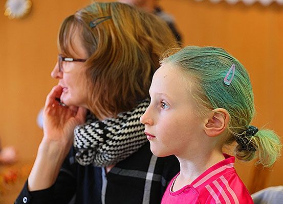 Eine Frau mit Brille und Schal schaut ein Mädchen mit grünem Haar an, beide sitzen in einem Raum.
