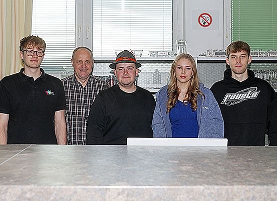 Fünf Personen stehen hinter einer Theke. Ein Mann mit Hut und ein anderer mit Brille stehen links, während ein Mann in einem karierten Hemd in der Mitte steht. Eine Frau in einem blauen Shirt und ein weiterer Mann in einem schwarzen Hoodie stehen rechts.