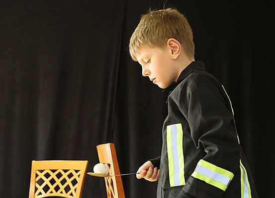 Ein Junge in einer Feuerwehruniform hält einen Löffel mit einer Kugel darauf, möglicherweise spielt er ein Spiel.