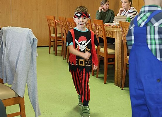 Ein junger Junge, der als Pirat verkleidet ist, geht in einem Raum mit Holzstühlen und Tischen. Er trägt einen Piratenhut, eine Bandana und Gesichtsbemalung. Ein weiteres Kind und zwei Erwachsene sitzen am Tisch im Hintergrund.
