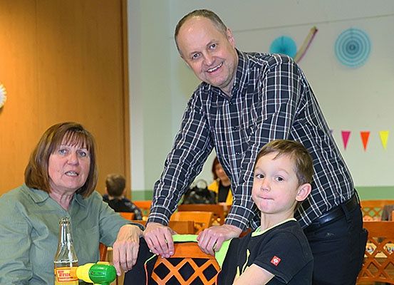 Eine Familie aus drei Personen, ein Mann, eine Frau und ein junger Junge, sitzen an einem Tisch, möglicherweise in einem Klassenzimmer, mit einer Flasche vor ihnen.