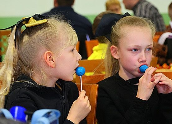 Zwei junge Mädchen mit blonden Haaren, die schwarze Pullover tragen, sitzen auf Stühlen und spielen mit blauen Lutschern. Sie scheinen sich in einem Klassenzimmer zu befinden.