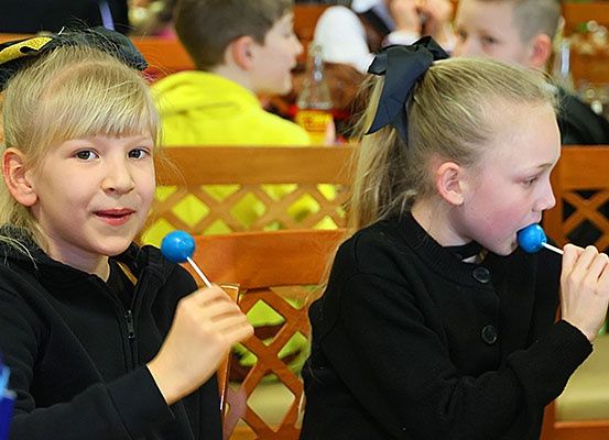Zwei junge Mädchen, gekleidet in Schwarz, sitzen an einem Tisch mit blauen Lutschern in den Händen. Sie befinden sich wahrscheinlich in einem Klassenzimmer. Hinter ihnen sitzt ein Junge in einem gelben Hemd, und weitere Kinder sind am Tisch platziert.
