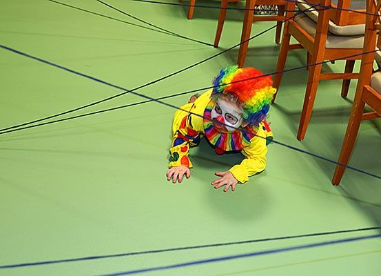 Ein Kind in einem Clownkostüm kriecht auf einem grünen Boden mit blauen Linien, umgeben von Holzstühlen.