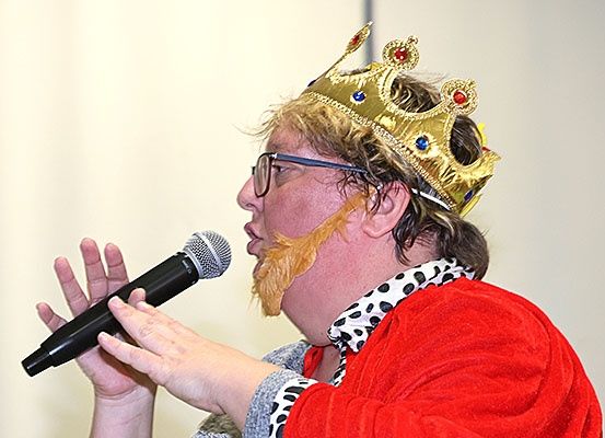 Eine Person mit Krone und rotem Umhang mit Bart und Schnurrbart hält ein Mikrofon.