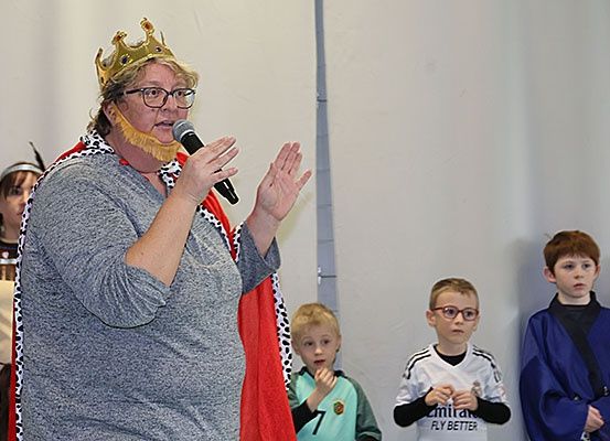 Eine Frau, die als König verkleidet ist, spricht in ein Mikrofon, während eine Gruppe von Kindern zusieht. Hinter ihr tragen zwei Kinder Kostüme.