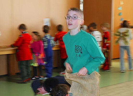 Ein Junge in einem grünen Shirt mit Brille hält einen Sack aus Jute, während andere hinter ihm stehen. Sie befinden sich in einem Raum mit grünem Boden und dekorativen Dreiecken an der Wand.