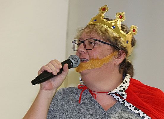 Eine Frau mit goldener Krone, rotem Umhang und falschem Bart hält ein Mikrofon. Sie sieht ernst aus und scheint aufzutreten.