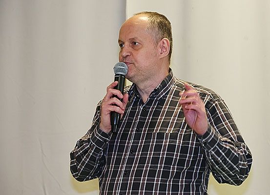 Ein Mann spricht in ein Mikrofon, das er mit beiden Händen hält, während er mit der anderen Hand gestikuliert. Er trägt ein kariertes Hemd.