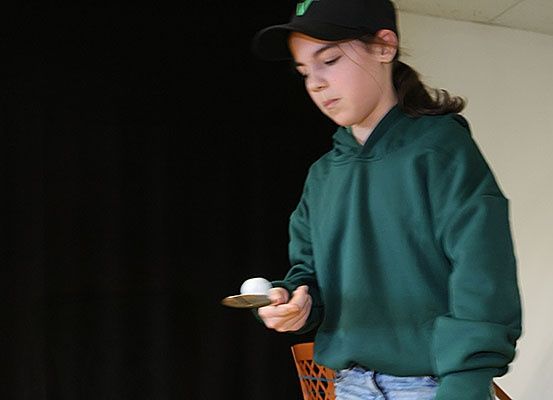 Ein junges Mädchen spielt Tischtennis. Sie trägt eine grüne Hoodie und eine schwarze Kappe. Sie hält einen Schläger und eine Kugel und schaut auf die Kugel. Sie steht in einem Raum mit einem schwarzen Vorhang und einer weißen Wand.
