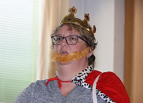 Eine Frau mit Krone, falschem Bart und rotem Umhang mit schwarzen Flecken steht in einem Raum.
