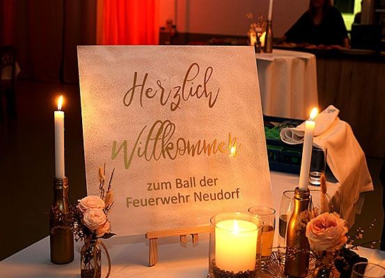 Ein Tisch mit einem Schild, das 'Herzlich Willkommen' steht. Es gibt Kerzen, Blumen und Gläser. Eine Person sitzt an einem anderen Tisch.