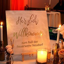 Ein Tisch mit einem Schild, das 'Herzlich Willkommen' steht. Es gibt Kerzen, Blumen und Gläser. Eine Person sitzt an einem anderen Tisch.