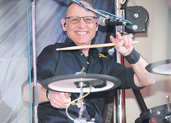 Ein Mann spielt auf der Bühne die Drums mit einem Mikrofon vor ihm. Er lächelt und hält Drumsticks.