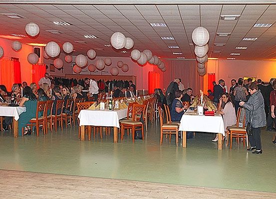Ein Bankettsaal mit gedeckten Tischen für eine formelle Veranstaltung. Die Leute sitzen und andere stehen. Der Raum ist mit Papierlaternen und roten Vorhängen dekoriert.