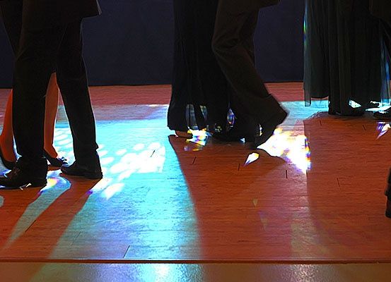 Eine Gruppe von Menschen tanzt auf einer Bühne mit blauen und weißen Lichtern, die Schatten auf den Boden werfen.