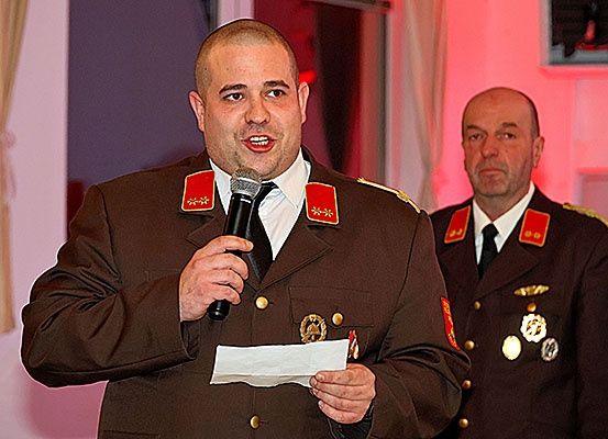 Ein Mann in Uniform hält ein Mikrofon und ein Papier in der Hand und spricht, während ein anderer Mann in Uniform hinter ihm steht.