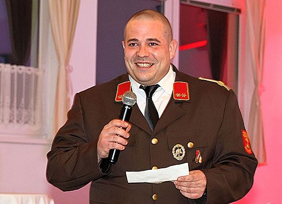 Ein Mann in Uniform, lächelt und hält ein Mikrofon und Papier in der Hand, spricht wahrscheinlich bei einer Veranstaltung.