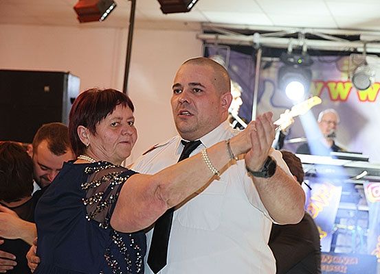 Eine ältere Frau in einem blauen Kleid tanzt mit einem Mann in einer weißen Uniform. Sie sind von anderen Leuten bei einer Party mit Bühne, Lautsprechern und Scheinwerfer umgeben.