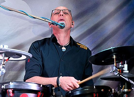 Ein Schlagzeuger spielt die Drums vor einem Mikrofon, trägt eine Brille und eine Halskette.