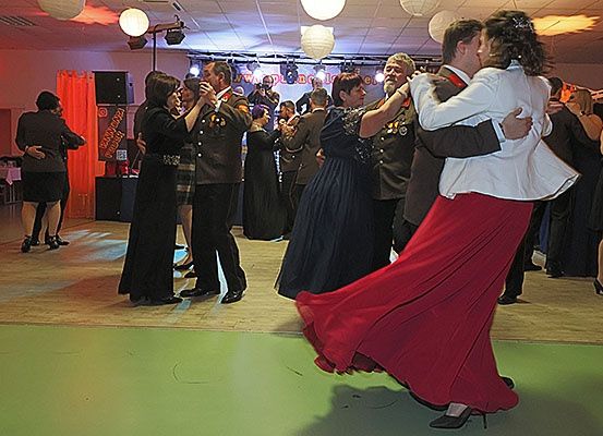 In einem schwach beleuchteten Raum tanzen Paare auf einem Holzboden. Eine Frau in einem roten Rock und ein Mann in einem weißen Blazer tanzen zusammen. Hinter ihnen tanzen andere Leute und einige stehen herum. Der Raum ist mit roten und weißen Lichtern und Vorhängen dekoriert.