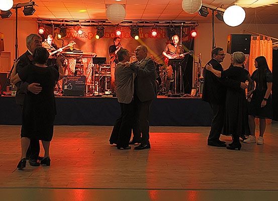 Eine Gruppe von Paaren tanzt auf einer Bühne, während eine Band hinter ihnen spielt. Die Bühne ist mit roten Lichtern beleuchtet.
