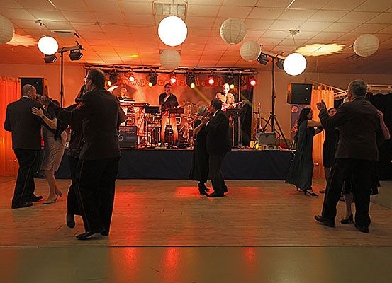 Eine Gruppe von Menschen in formeller Kleidung tanzt auf einer Tanzfläche. Eine Band spielt auf einer Bühne mit Musikinstrumenten. Der Bereich ist mit roten Lichtern beleuchtet.